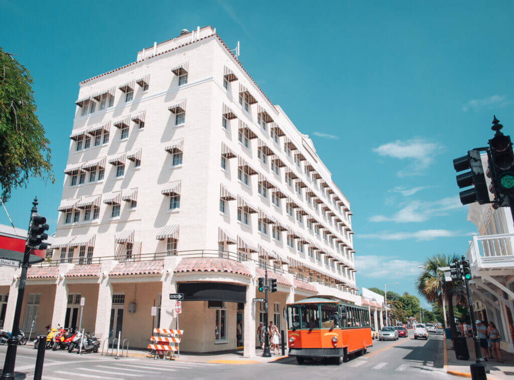 Photo taken on a sunny day of Duval Street, in Key West. An orange old town trolley tour vehicle is driving down the street.
